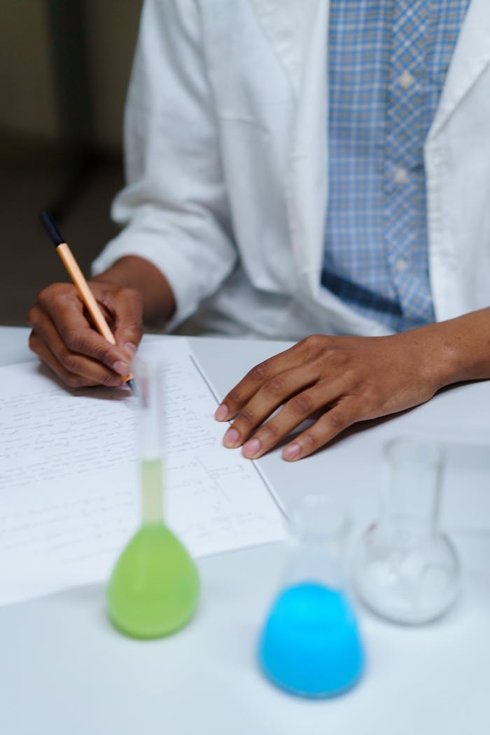 why-choose-us A student in a lab coat writes notes beside flasks filled with colored liquids in a laboratory setting.